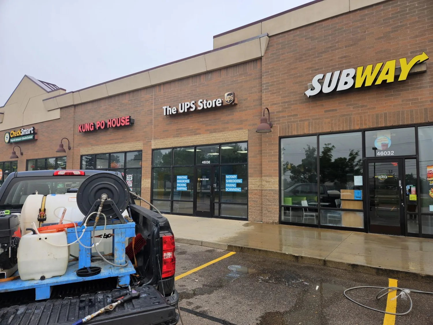 A multi-tenant retail plaza with the walkway and entrance aprons freshly washed — the concrete is still wet — and their pressure-wash rig, tank and hose reel sitting in the bed of the truck in the foreground.